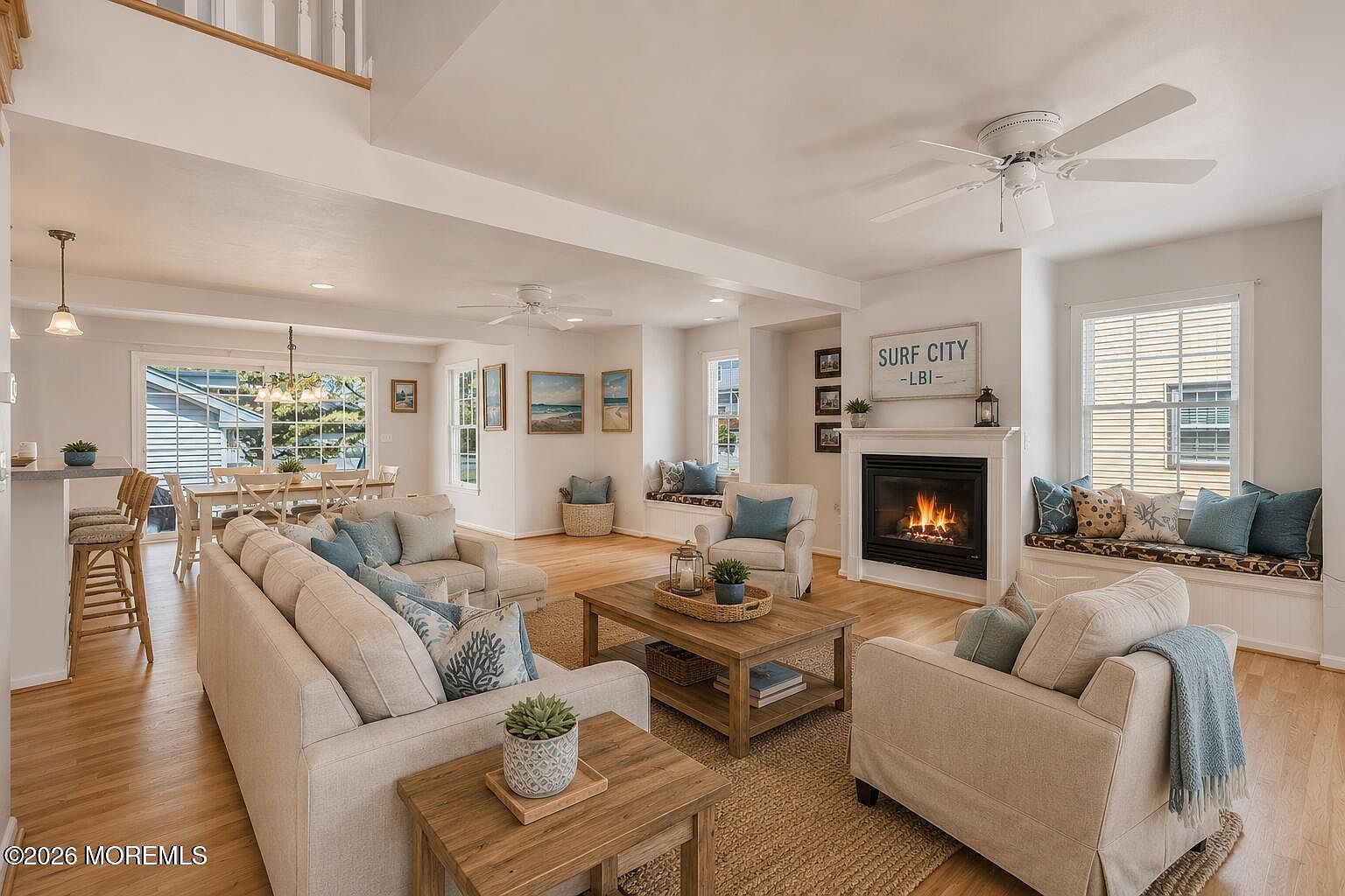 The great room at 233 N 7th Street fully furnished in coastal style, with sofas and armchairs around the gas fireplace, the SURF CITY LBI sign on the mantel, window-seat benches with blue cushions, and the kitchen visible through the open archway